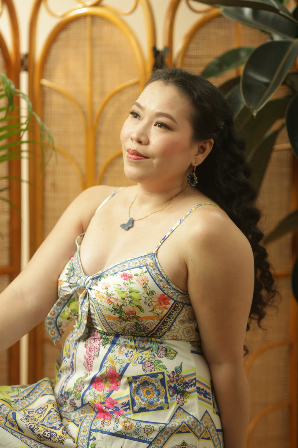 Woman wearing a colorful patterned dress sitting against a wooden screen with plants.