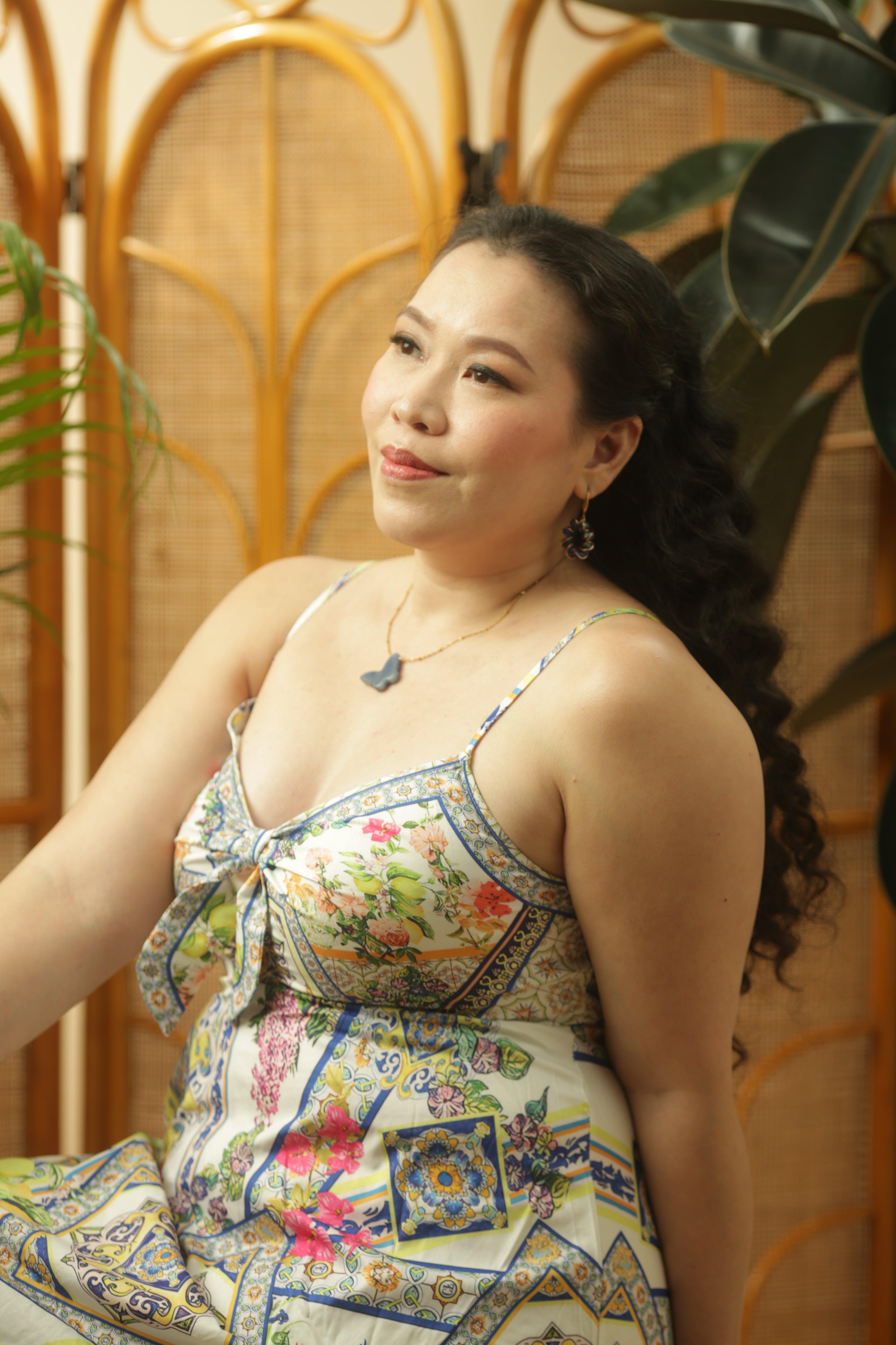 Woman wearing a colorful patterned dress sitting against a wooden screen with plants.