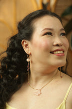 Woman with long dark hair and earrings, wearing a yellow top, against a blurred background.